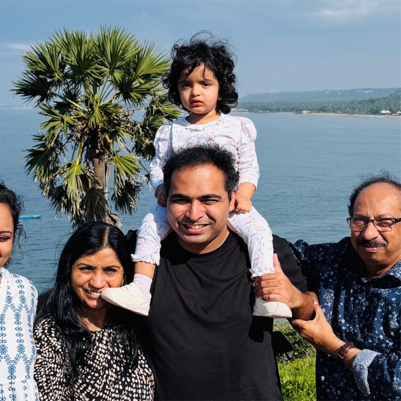 Akhil Suresh Nair smiling with his family by the sea, a young child sitting on his shoulders.